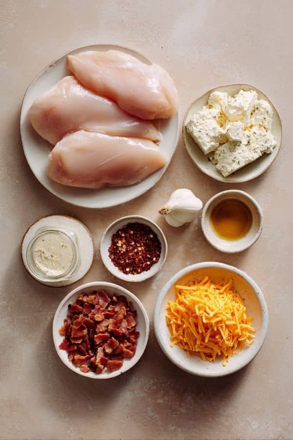 Ingredients for Crockpot crack chicken including chicken breasts, ranch seasoning, cream cheese, cheddar cheese, and bacon arranged on a countertop
